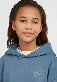 Sudadera con capucha azul con un diseño de corazón metálico en el pecho. Textura suave, corte relajado, usada sobre una camiseta blanca.