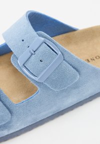 Blå mockaskandal med justerbart rem och fyrkantig spänne, med en brun texturerad fotbädd och öppen sidodesign för andningsförmåga.