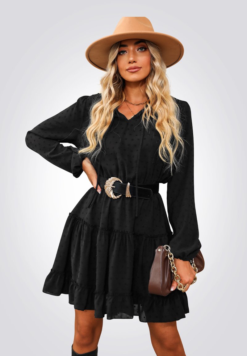 Vestido negro con lunares, de mangas largas y con un dobladillo voluminoso, ceñido a la cintura con un cinturón con detalles dorados. Combinado con un bolso marrón y un sombrero color beige.