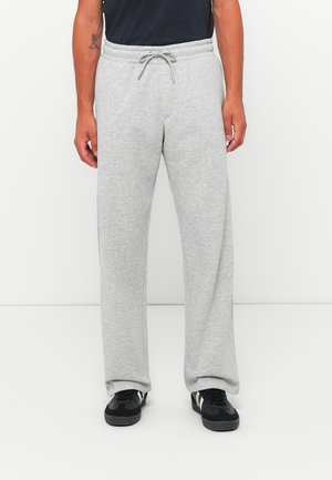 Uomo in pantaloni da jogging grigi chiari con cintura regolabile e scarpe nere con striscia bianca, in piedi contro uno sfondo bianco.