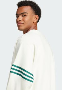 Vit sweatshirt med rund hals och gröna ränder på ärmarna. Tyget verkar mjukt med en texturerad yta.