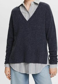 Marineblauer Pullover mit V-Ausschnitt, geripptem Saum und langen Ärmeln über einem gestreiften Hemd mit Kragen. Der Stoff wirkt weich und strukturiert.