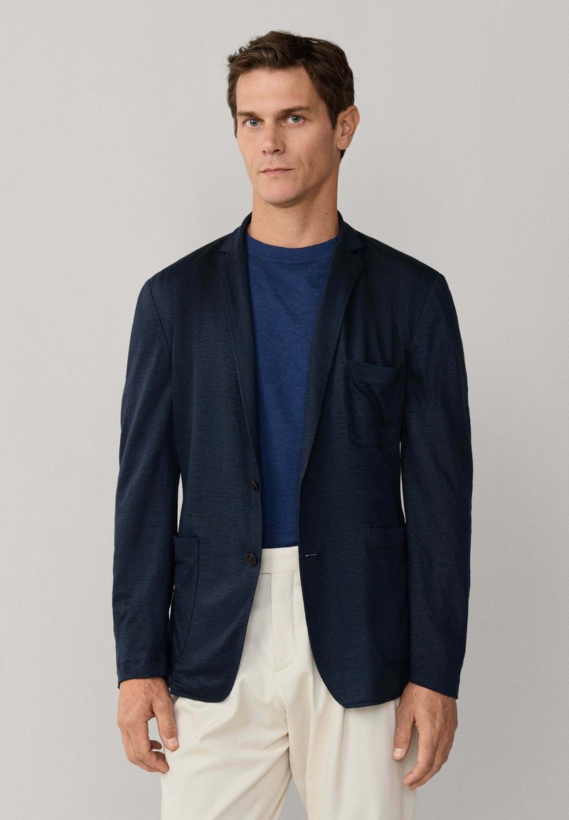 Mladý muž v modrom saku cez modrú košeľu a krémové nohavice, stojaci pred jednoduchým svetlošedým pozadím.