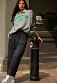 Sivkasta oversized pulover sredi zelenega napisa "NEW YORK 1986", v kombinaciji s temnimi širokimi hlačami. Model se naslanja na črno betonsko odbojnico.