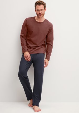 Man draagt een bruin shirt met lange mouwen en donkergrijze broek, blootsvoets, staand en leunend tegen een lichtgekleurde muur.