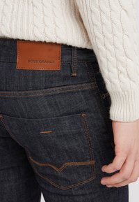 Mörka denimjeans med orange sömmar, som har en brun läderpatch märkt "BOSS ORANGE" och en formad bakfickdesign.