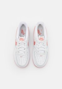 Pár bílých tenisek Nike Air Force 1 s růžovým swooshem a akcenty, zavázaných a zobrazených z vrchu na bílém pozadí.