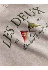 Felpa grigia con testo ricamato e motivi di bandiera in verde, rosso e giallo. Dettagli notevoli includono "LES DEUX" e "EST. 2011".