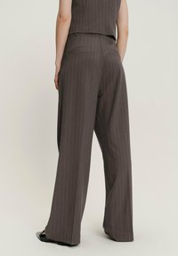 Mujer vistiendo pantalones de pinzas marrones de cintura alta y pernera ancha, junto con una blusa sin mangas a juego, de pie frente a un fondo liso.