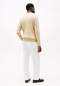 Μπεζ πουλόβερ με καλωδιακό πλέξιμο και ριμπ τελείωμα, φορεμένο με λευκό παντελόνι και σκούρα παπούτσια· παρουσιάζει υφασμένη όψη από πίσω.