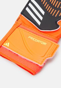 Guanto da portiere arancione con presa in silicone testurizzato, accenti neri, strisce bianche e logo "PREDATOR" dorato sul cinturino del polso.