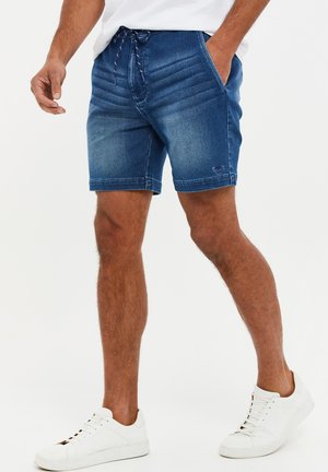 Homme portant un short en denim bleu avec des poches latérales et des baskets blanches, se tenant avec une main dans sa poche sur fond blanc.