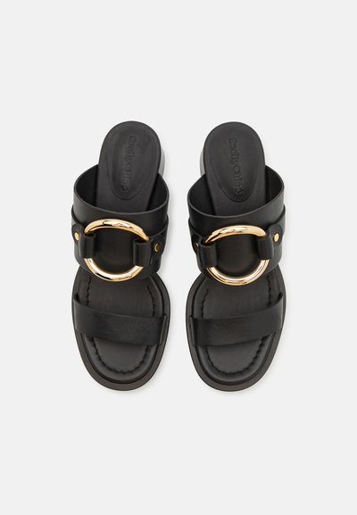 Crne klizne sandals sa širokim remenom, izrađene od glatke kože, s zlatnim okruglim detaljem i podstavljenom unutrašnjošću za udobnost.