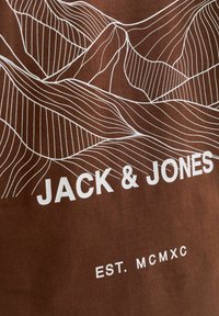 T-shirt marron avec des graphismes en ligne de montagne blanche et le texte "JACK & JONES", avec "EST. MCMXC" en dessous. Tissu en coton texturé.