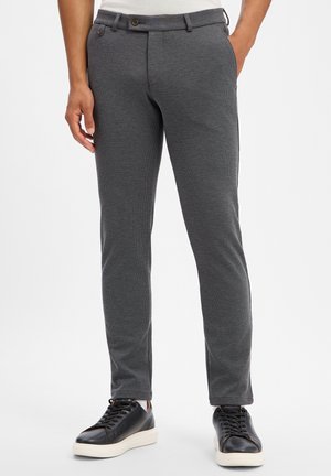 Man draagt een slim-fit donkergrijze broek en zwarte sneakers met witte zolen, staand met één hand in de zak tegen een witte achtergrond.