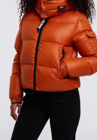 Pomarańczowa puchowa kurtka z wysokim kołnierzem, zamkiem z przodu, bocznymi kieszeniami i elastycznymi mankietami. Błyszcząca faktura z pikowanymi panelami zapewniającymi izolację.