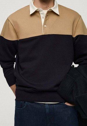 Homme portant un polo à manches longues bicolore avec une partie supérieure beige, une partie inférieure noire, un col blanc et un jean bleu, tenant une veste sombre.