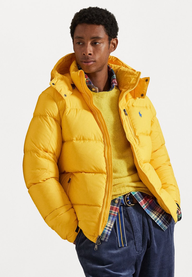 Yellow padded jacket with a zip front, attached hood, and logo. Layered over a yellow sweater and plaid shirt. Textured fabric.
