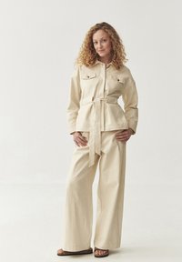 Beige puuvillane komplekt, millel on vööga jakk kahte rinnataskut ja laiad püksid. Mudel seisab ühe käega vööl.