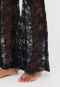 Pantaloni in pizzo nero con un motivo floreale. Materiale trasparente che mostra la pelle sottostante. L'orlo presenta un bordo a onde. I piedi scoperti sono visibili sotto.