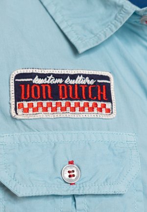 Tissu bleu clair avec patch brodé "Von Dutch" en motif damier rouge et blanc au-dessus d'une poche boutonnée avec un bouton de marque.