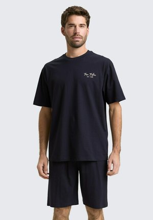 Man draagt een oversized zwart t-shirt met de tekst "Tom Tailor EST. 1962" en bijpassende zwarte knielange shorts, staand tegen een effen achtergrond.