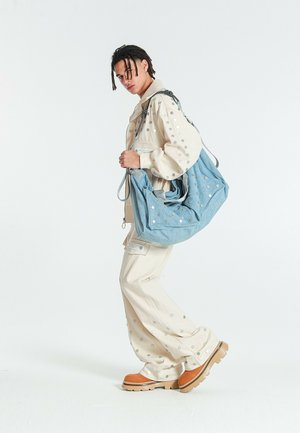 Joven con cabello trenzado lleva un conjunto beige con puntos metálicos, lleva un gran bolso de hombro azul con tachuelas y botas marrones.