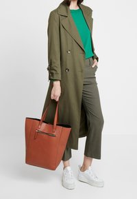 Olijfgroene lange jas, groene top, olijfgroene broek, witte sneakers. Oranje draagtas met gestructureerd oppervlak en ritszak.