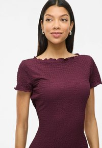 Vestido carmesí con un acabado texturizado, cortas mangas con volantes y un escote con ondas, diseño ajustado que acentúa la silueta.