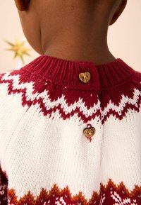 Le pull en tricot présente un motif en chevrons bordeaux et blanc avec des boutons en forme de cœur en bois à l'arrière et un col côtelé.