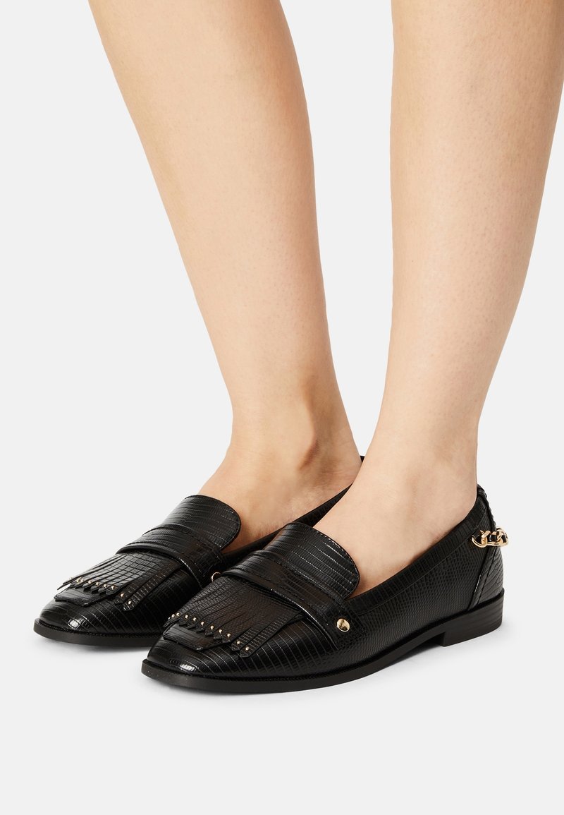 Mocassins en cuir texturé noir avec franges et détails cloutés, portés pieds nus sur un fond blanc.