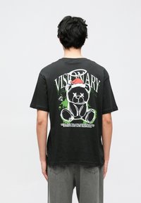 Tricou negru din bumbac, cu un desen grafic al unui urs cu o căciulă de Moș Crăciun, textul "VISIONARY" și accente verzi, cu croială lejeră, supradimensionată.