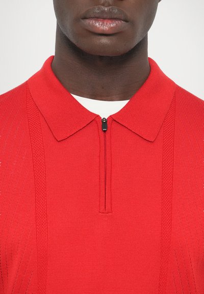 Punainen neulottu poolopaita vetoketjullisella kauluksella, jossa on teksturoidut raidat ja sileä pinta. Yhdistetty valkoisen vaatteen kanssa.