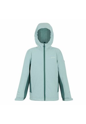 Lachend meisje met lang blond haar, gekleed in een lichtblauw jasje, crèmekleurige broek, bruine enkellaarzen en een grijze crossbodytas, staat tegen een effen achtergrond.