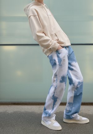 Felpa beige con tasca frontale e cappuccio, abbinata a pantaloni con motivo nuvole blu e bianche. Il modello indossa sneakers bianche e grigie.