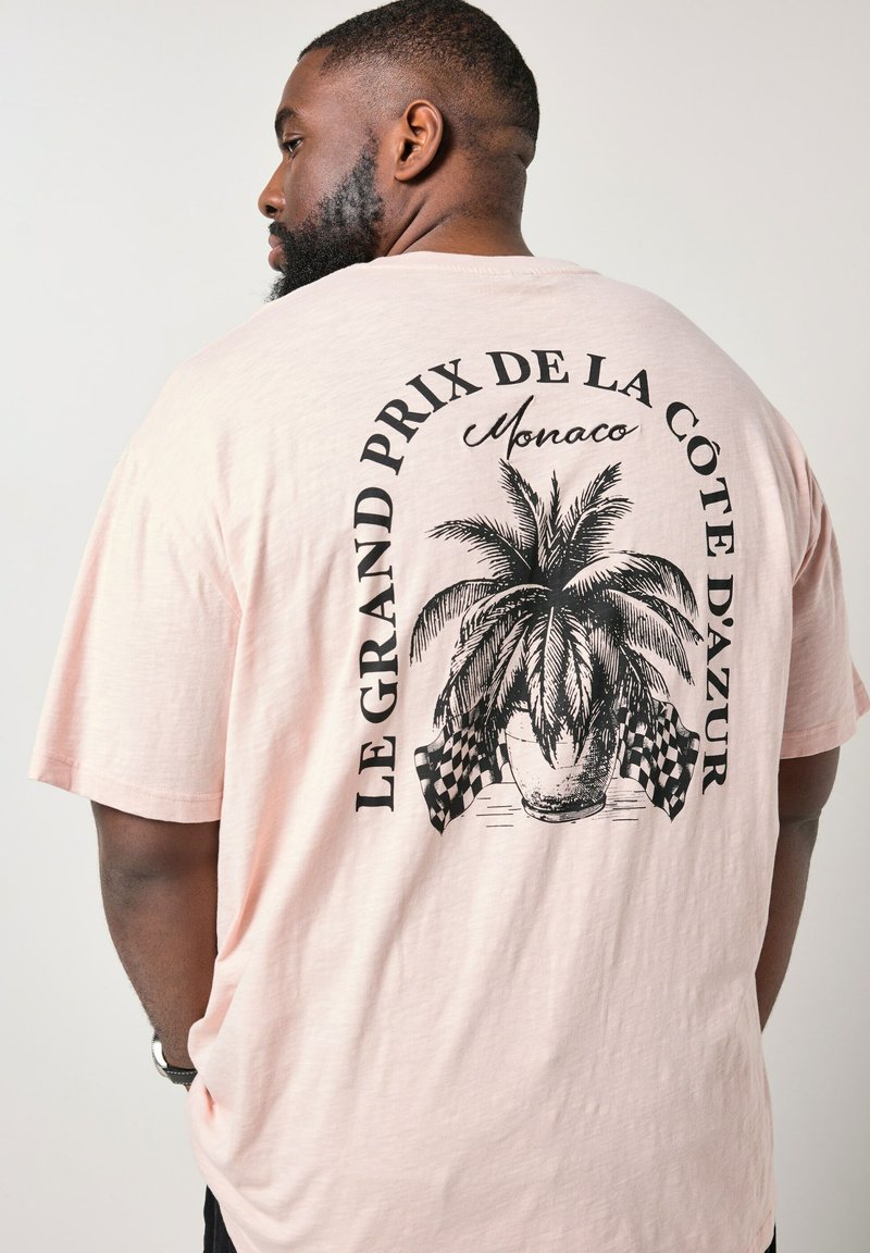 Mand iført lyserød t-shirt med sort print af en palme i potte og ternede flag, tekst "Le Grand Prix de la Côte d'Azur Monaco" på ryggen.