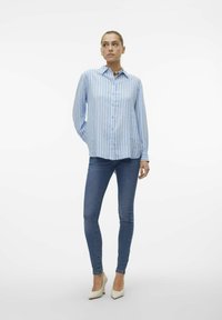 Light blue long-sleeve shirt with white and blue vertical stripes, button-down front, paired with fitted blue jeans and pointed white heels.