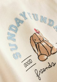 Der cremefarbene Stoff zeigt gestickte Texte "SONNTAG" und "GUTE FREUNDE" mit einem gedruckten braunen Hasenmotiv, einschließlich Akzenten und einem roten Band.