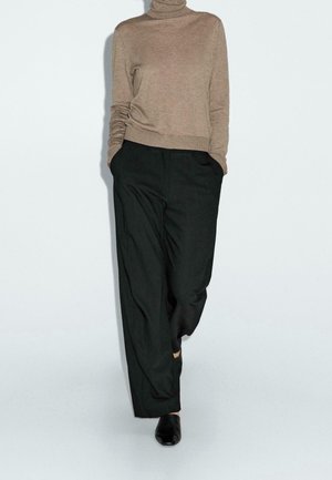 Personne portant un pull col roulé beige, un pantalon noir ample avec les mains dans les poches, et des chaussures noires à bouts pointus sur un fond uni.