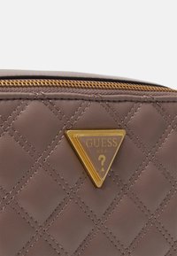 Gequilte taupe leren tas met een ritssluiting en een gouden driehoekig Guess USA-logo met een vraagteken in het midden.