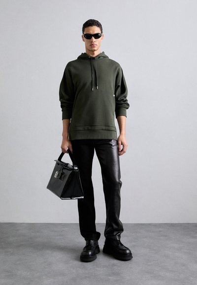Dunkelgrüner Kapuzenpullover mit einer Fronttasche, schwarze Kunstlederhose und schwarze Plateauschuhe. Halten eine schwarze strukturierte Handtasche mit Hardware.