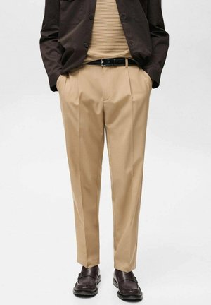 Man draagt beige geplooide broek, bruine leren riem, donkerbruine jas, beige gebreide top en donkerbruine leren loafers met handen in de zakken.