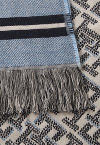 Geweven blauw en marine gestreepte textiel met zwart-witte franje, laag over een patroon van lichte stof met geometrische ontwerpen.