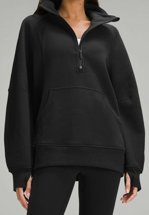 Femme portant un sweat à capuche noir oversize avec fermeture éclair au quart et poche kangourou à l'avant, debout devant un fond gris uni.