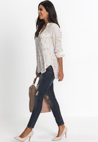 Blusa floreale leggera con maniche lunghe e orlo asimmetrico, abbinata a jeans scuri strappati e tacchi nudi, mentre tiene una borsa beige.