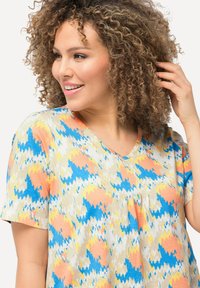 Top de cuello en V en tejido suave con un patrón de zigzag multicolor en azul, naranja y amarillo sobre un fondo beige; mangas cortas.