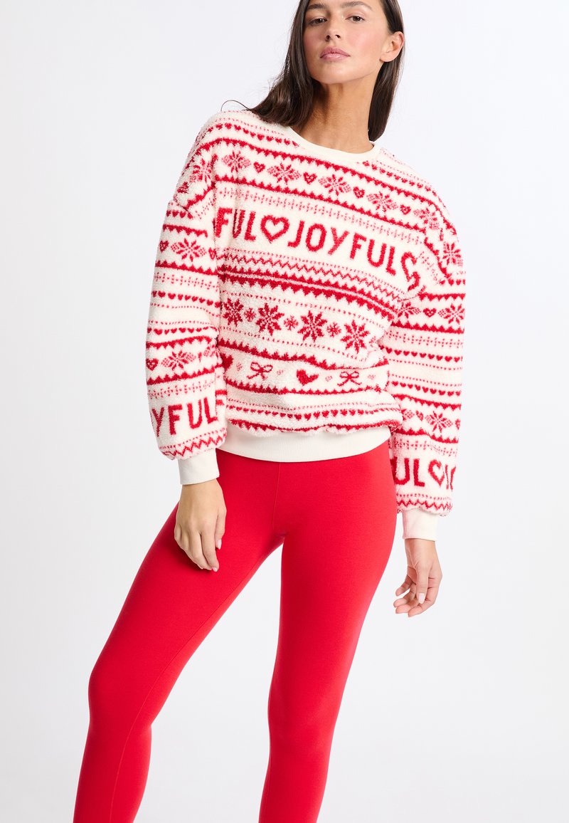 Czerwony i biały sweter z dzianiny z wzorami serc i płatków śniegu, okrągłym dekoltem i ściągaczami na mankietach. Połączony z dopasowanymi czerwonymi legginsami.