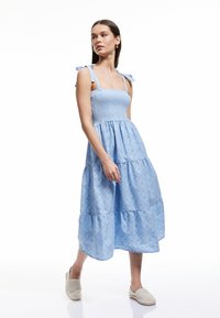 Vestido azul claro con corsé fruncido y falda acampanada, que presenta tirantes en los hombros y un patrón de ojales. Combinado con zapatos de slip-on en tonos neutros.