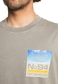 Šedé bavlněné tričko s kulatým výstřihem, na levé straně se modro-oranžovým logem. Jemná textura s lehkými značeními.