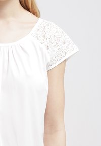 Blouse blanche à manches courtes, avec détails en dentelle sur les épaules, tissu doux et encolure froncée pour une coupe décontractée.